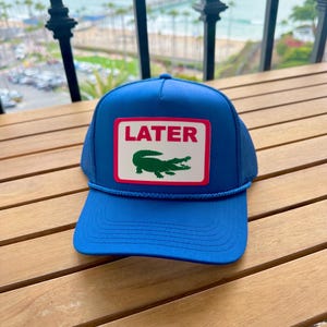 May include: A blue trucker hat with a white rectangle patch featuring the word "LATER" in red and a green alligator logo. The hat has a mesh back and a blue rope detail.