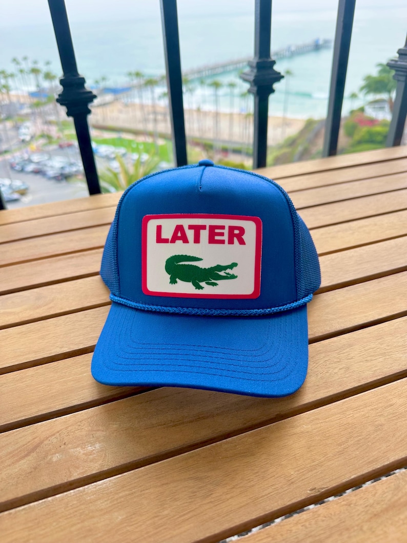 May include: A blue trucker hat with a white rectangle patch featuring the word "LATER" in red and a green alligator logo. The hat has a mesh back and a blue rope detail.