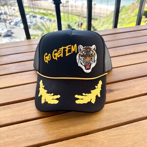 May include: Black trucker hat with a yellow brim and a tiger face patch. The hat also has a yellow embroidered design on the front that says "Go Get Em".
