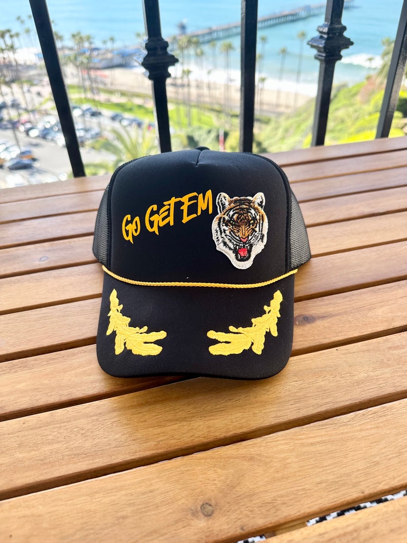 May include: Black trucker hat with a yellow brim and a tiger face patch. The hat also has a yellow embroidered design on the front that says "Go Get Em".