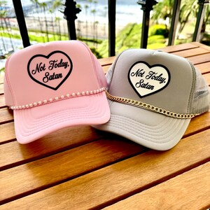 May include: Two trucker hats with a heart-shaped patch that says "Not Today, Satan". One hat is pink with a pearl trim and the other is gray with a gold chain.