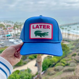 May include: A blue trucker hat with a white and red rectangular patch that reads "LATER" above a green alligator logo. The hat has a blue brim and mesh backing. A blue rope detail accents the brim.