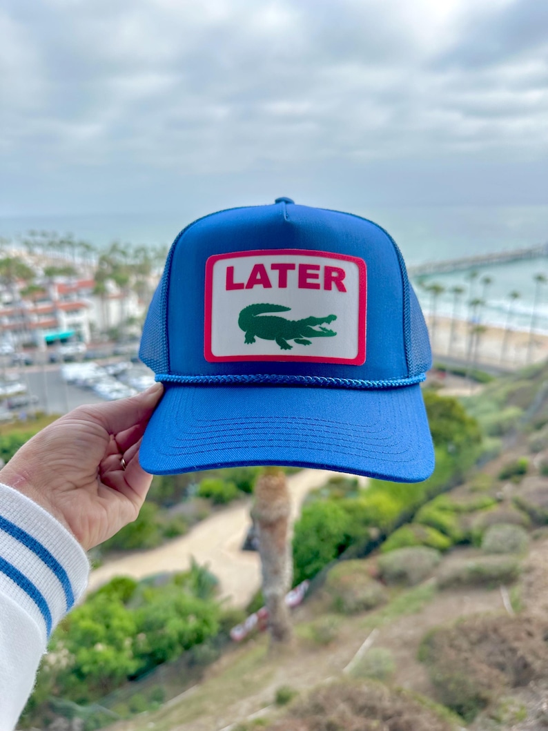 May include: A blue trucker hat with a white and red rectangular patch that reads "LATER" above a green alligator logo. The hat has a blue brim and mesh backing. A blue rope detail accents the brim.