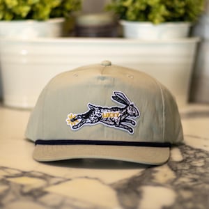 Puede incluir: Gorra de béisbol beige con un detalle de cuerda azul marino y un parche con un conejo corriendo con la palabra "LUCKY". El conejo es blanco y negro con detalles amarillos. La gorra está sobre una superficie de mármol.