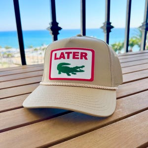 May include: Tan trucker hat with a white and red rectangular patch featuring the word "LATER" above a green alligator. The hat has a mesh back and a rope detail along the brim.