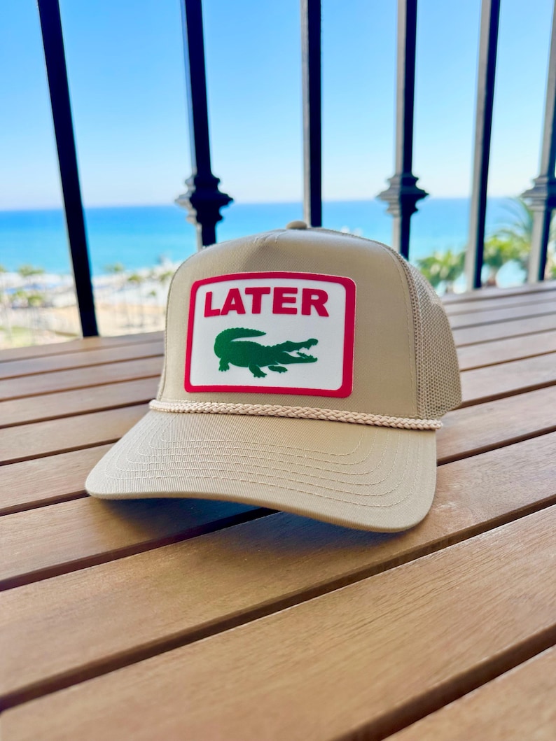 May include: Tan trucker hat with a white and red rectangular patch featuring the word "LATER" above a green alligator. The hat has a mesh back and a rope detail along the brim.
