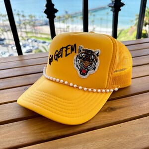 May include: A yellow trucker hat with a black embroidered tiger and the text "Go Get Em". The hat has a white pearl trim.