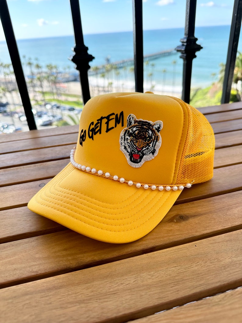 May include: A yellow trucker hat with a black embroidered tiger and the text "Go Get Em". The hat has a white pearl trim.