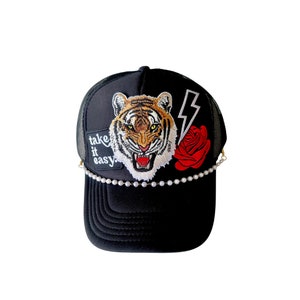 Könnte beinhalten: Schwarze Trucker-Kappe mit einem gestickten Tiger, einer Rose und einem Blitz auf der Vorderseite. Die Kappe hat außerdem einen Perlensaum und den Text "Take it easy" an der Seite.