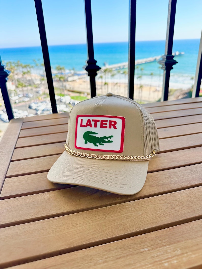 May include: Tan trucker hat with a mesh back and a curved brim. The front patch features the word "LATER" in red, with a green crocodile logo. A gold chain detail accents the brim.