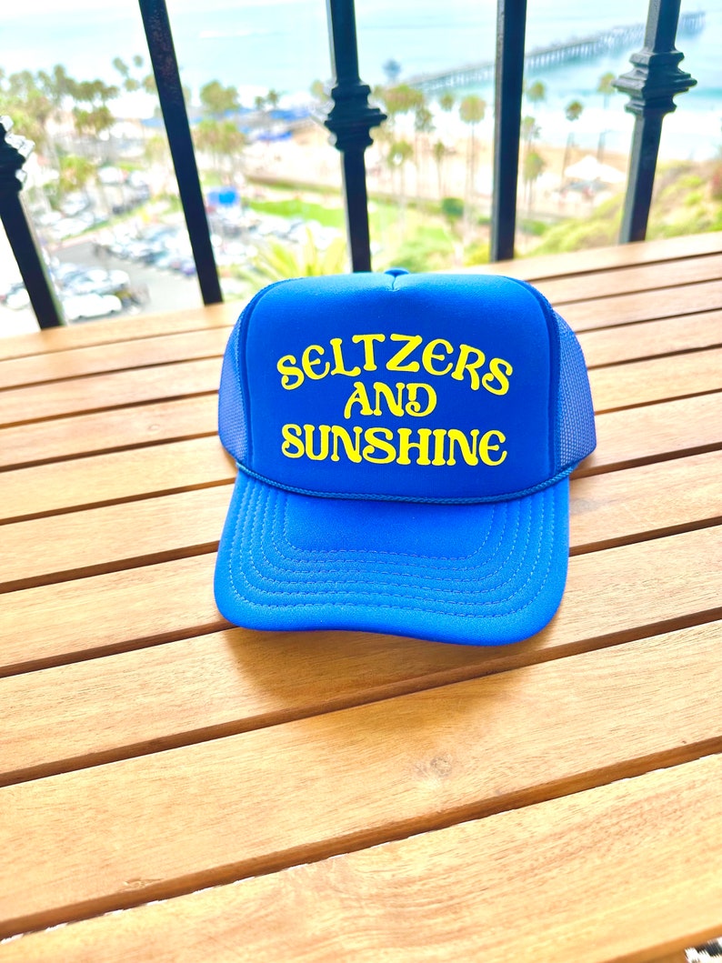 May include: A blue trucker hat with a yellow embroidered design that says "Seltzers and Sunshine".
