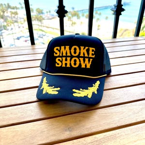 May include: Black trucker hat with a yellow mesh back. The front of the hat has the words "SMOKE SHOW" embroidered in yellow.