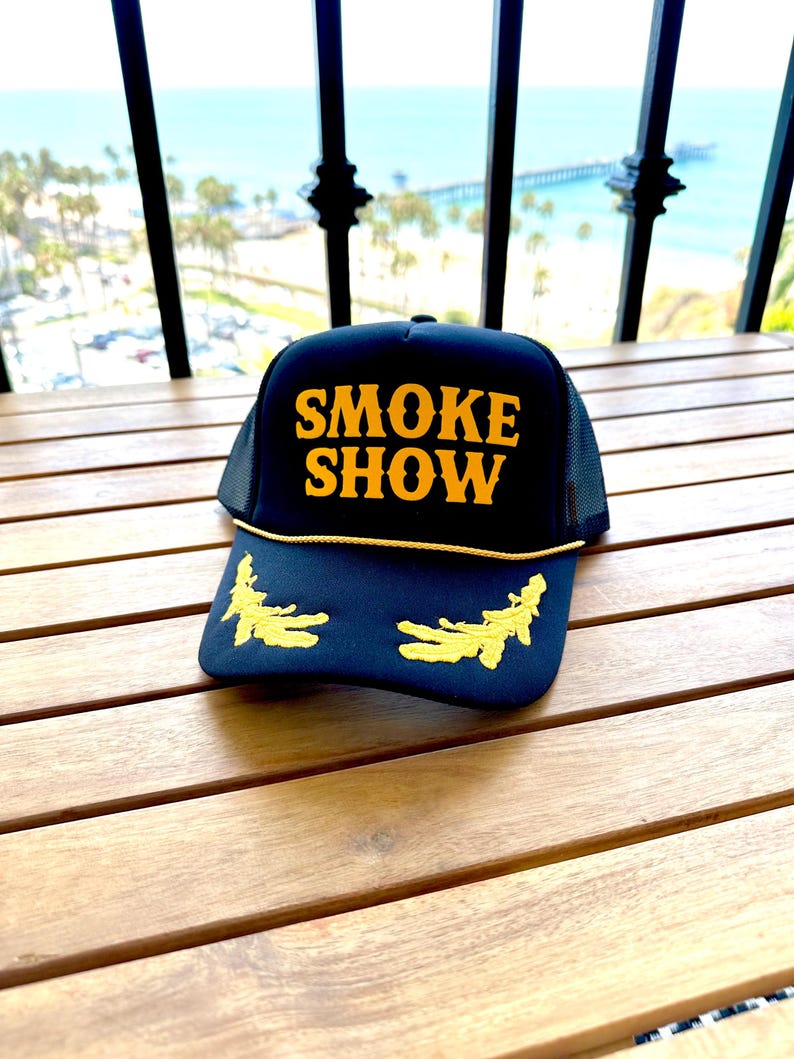 May include: Black trucker hat with a yellow mesh back. The front of the hat has the words "SMOKE SHOW" embroidered in yellow.