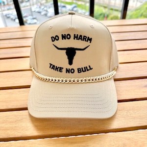 May include: A beige trucker hat with a gold chain around the brim. The hat has black embroidery that reads "Do No Harm Take No Bull" and features a bull skull graphic.