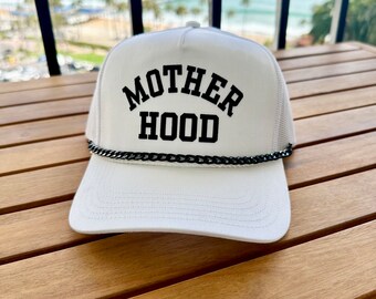 Pode incluir: Boné de caminhoneiro branco com acabamento de corrente preta e o texto "MOTHERHOOD" em letras maiúsculas pretas.