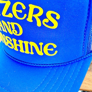 May include: Blue and yellow trucker hat with the text "Sizzlers and Sunshine" embroidered on the front.