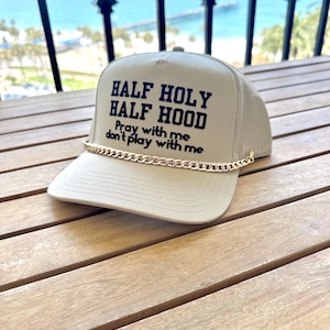 Puede incluir: Una gorra tipo trucker de color beige con parte trasera de malla y una cadena dorada sobre la visera. El panel frontal muestra el texto negro "HALF HOLY HALF HOOD" en negrita, seguido del texto negro más pequeño "Pray with me don't play with me."