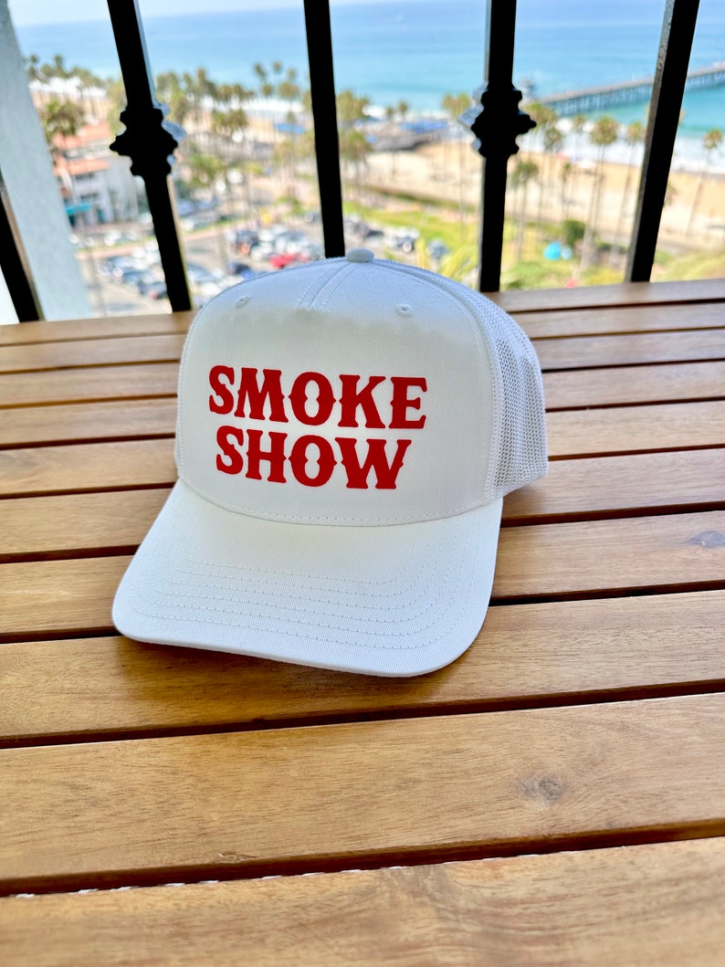 May include: White mesh trucker hat with red embroidered text that reads "SMOKE SHOW".