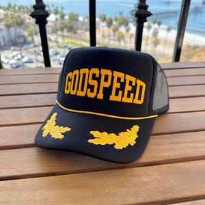 May include: Black trucker hat with a yellow "GODSPEED" embroidered on the front and two yellow embroidered oak leaf designs on the brim.