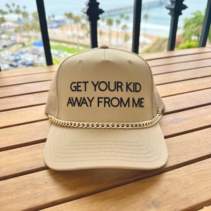 Könnte beinhalten: Ein beiger Trucker-Hut mit einer goldenen Kette um den Rand. Der Hut hat einen schwarzen gestickten Text mit der Aufschrift "GET YOUR KID AWAY FROM ME."