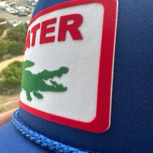 May include: Blue trucker hat with a white patch featuring the word "ATER" in red and a green alligator logo. The hat has a blue rope detail and a mesh back.
