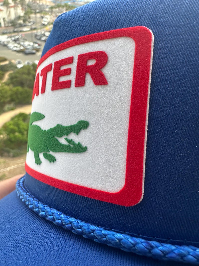 May include: Blue trucker hat with a white patch featuring the word "ATER" in red and a green alligator logo. The hat has a blue rope detail and a mesh back.
