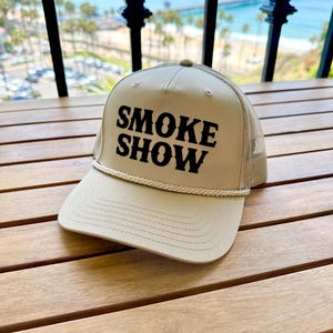 May include: A beige trucker hat with a black embroidered logo that says "SMOKE SHOW".