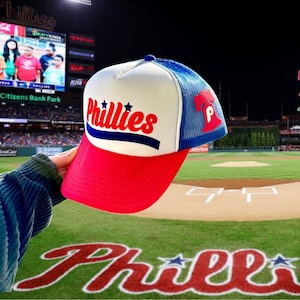 May include: A white and blue baseball cap with a red brim. The cap features a red, white, and blue "Phillies" logo on the front and a blue Liberty Bell logo on the side. The cap is resting on a baseball field with the word "Phillies" written in red on the grass.