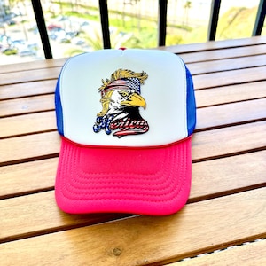 Puede incluir: Gorra de camionero con un panel frontal blanco con un gráfico de águila con una cinta patriótica y la palabra "America". La gorra tiene una visera rosa, laterales de malla azul y una parte superior roja.