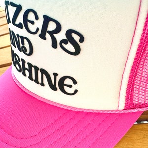 May include: A pink and white trucker hat with the text "Sizzlers and Shine" in black.
