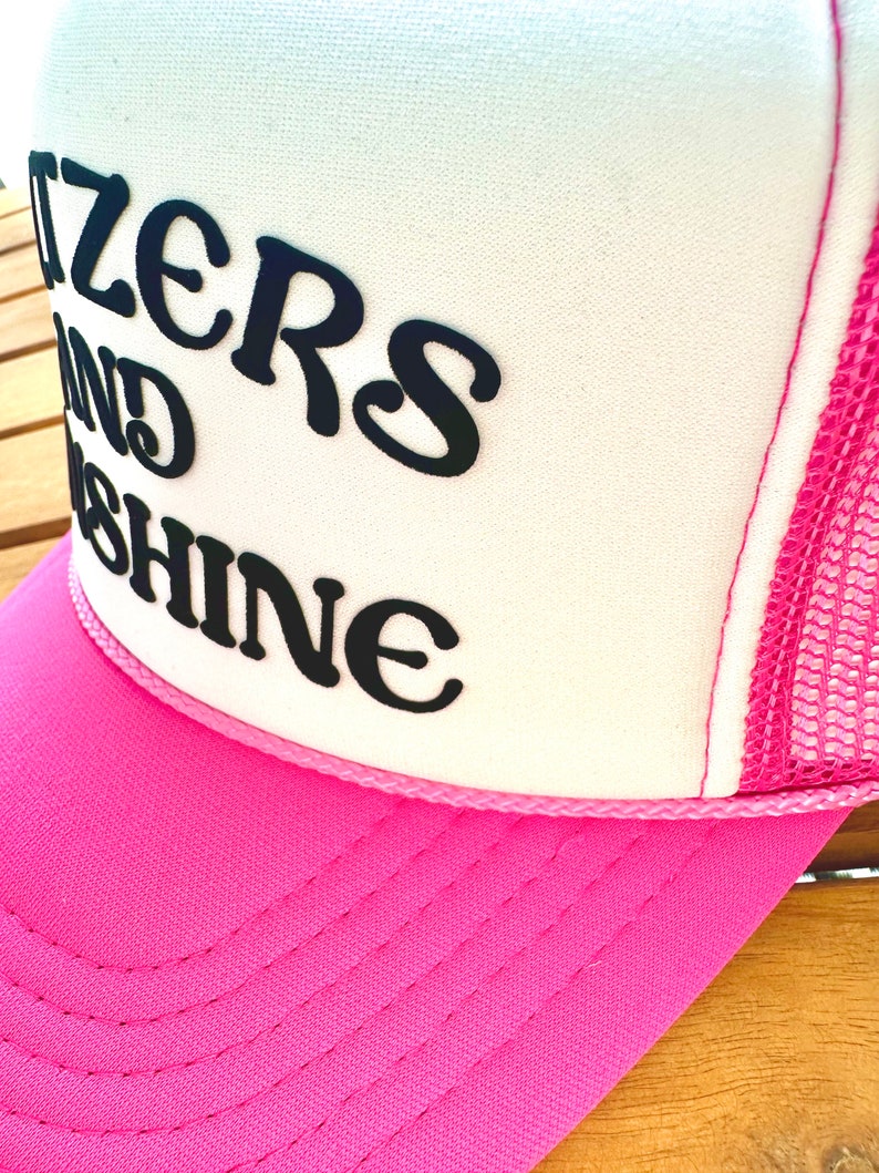 May include: A pink and white trucker hat with the text "Sizzlers and Shine" in black.