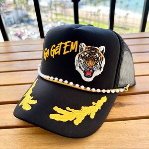 May include: Black trucker hat with a yellow embroidered tiger and the words "Go Get Em" in yellow. The hat has a white pearl chain around the brim.