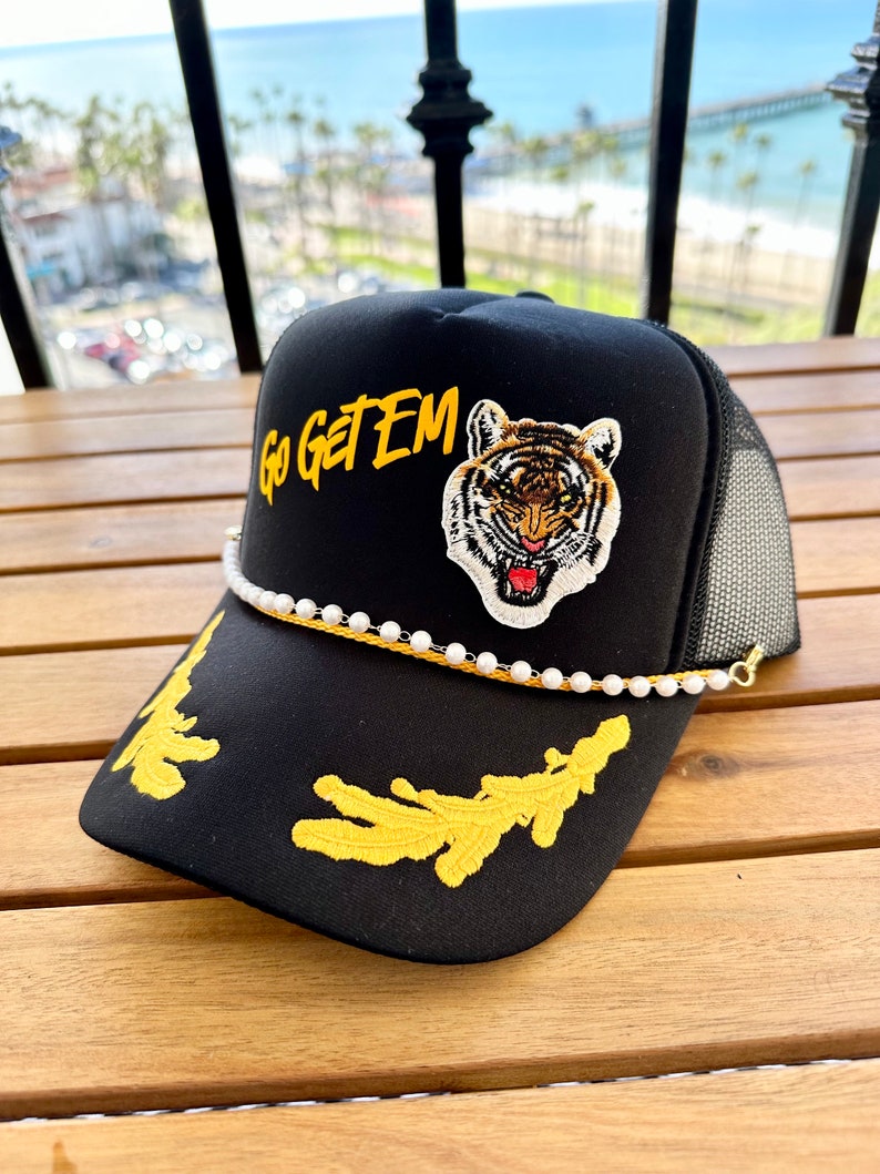 May include: Black trucker hat with a yellow embroidered tiger and the words "Go Get Em" in yellow. The hat has a white pearl chain around the brim.