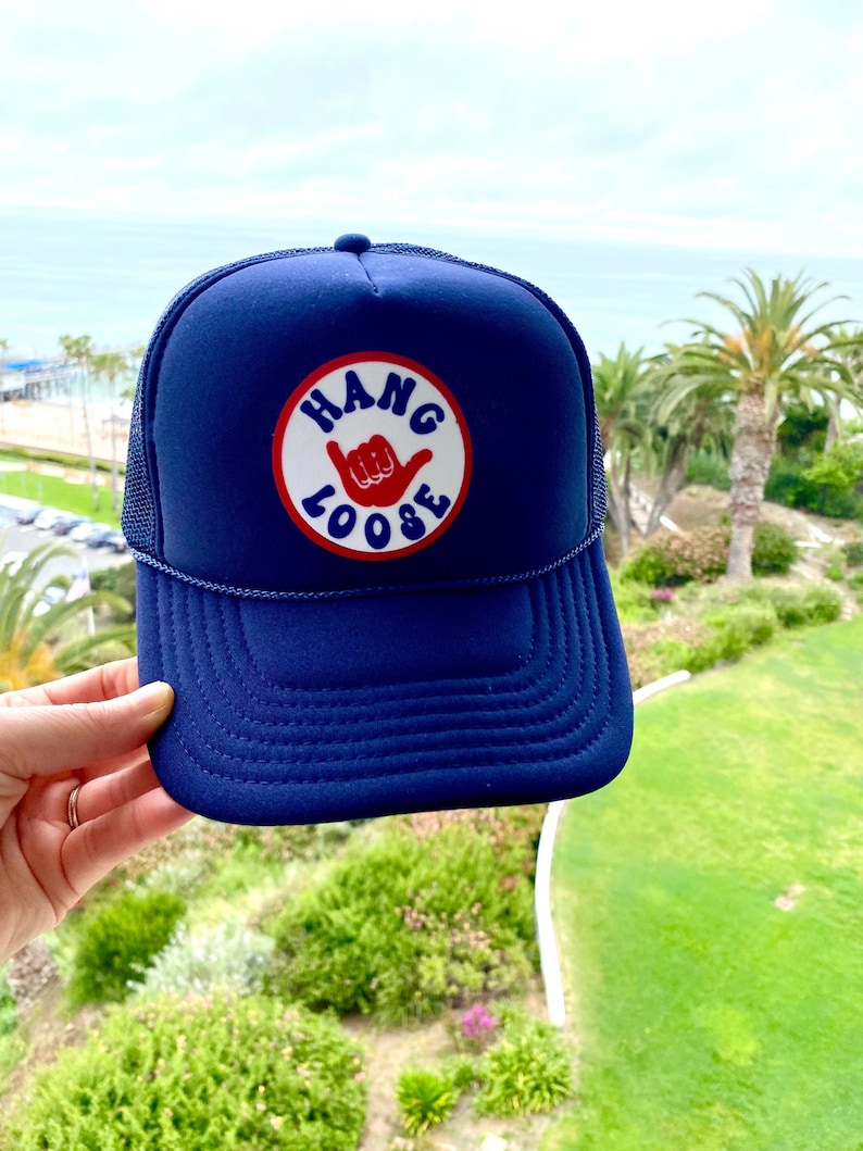 K&ouml;nnte beinhalten: Marineblaue Trucker-Cap mit einem wei&szlig;en und roten runden Aufn&auml;her mit der Aufschrift "HANG LOOSE" und einer Handgeste. Die Kappe hat einen Mesh-R&uuml;cken und einen gebogenen Schirm. Im Hintergrund ist eine K&uuml;stenlandschaft zu sehen.