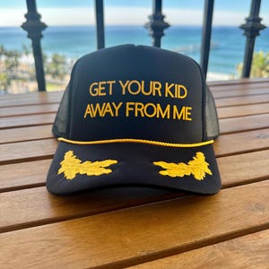 May include: Black trucker hat with a yellow embroidered brim and two yellow embroidered laurel wreaths on the front. The hat has the text "GET YOUR KID AWAY FROM ME" embroidered in yellow on the front.