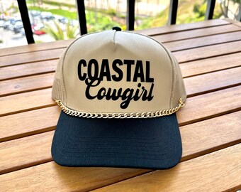 Könnte beinhalten: Eine beige und schwarze Trucker-Kappe mit goldener Kettenverzierung. Die Kappe hat den Schriftzug "COASTAL Cowgirl" in Schwarz auf der Vorderseite gestickt.