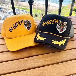 Puede incluir: Dos gorras de camionero con un gráfico de tigre y el texto "Go Get'em" bordado en la parte delantera. Una gorra es amarilla y la otra es negra.