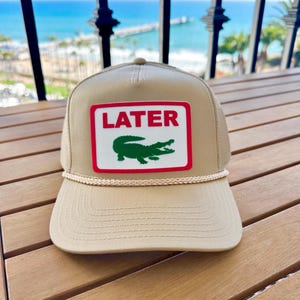May include: Tan trucker hat with a white and red patch featuring the word "LATER" above a green alligator logo. The hat has a curved brim and a rope detail. The background shows a view of the ocean.