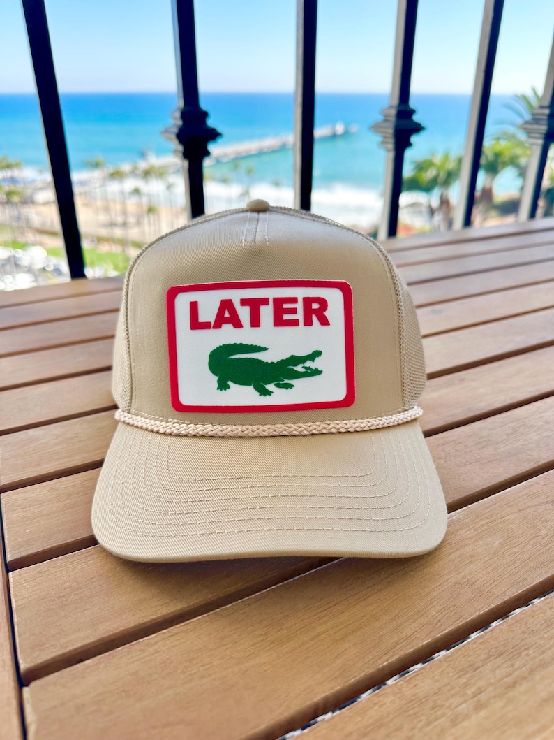 May include: Tan trucker hat with a white and red patch featuring the word "LATER" above a green alligator logo. The hat has a curved brim and a rope detail. The background shows a view of the ocean.