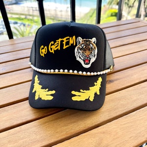 May include: Black trucker hat with a gold embroidered tiger and the words "Go Get Em" in yellow. The hat has a white pearl trim and a black mesh back.