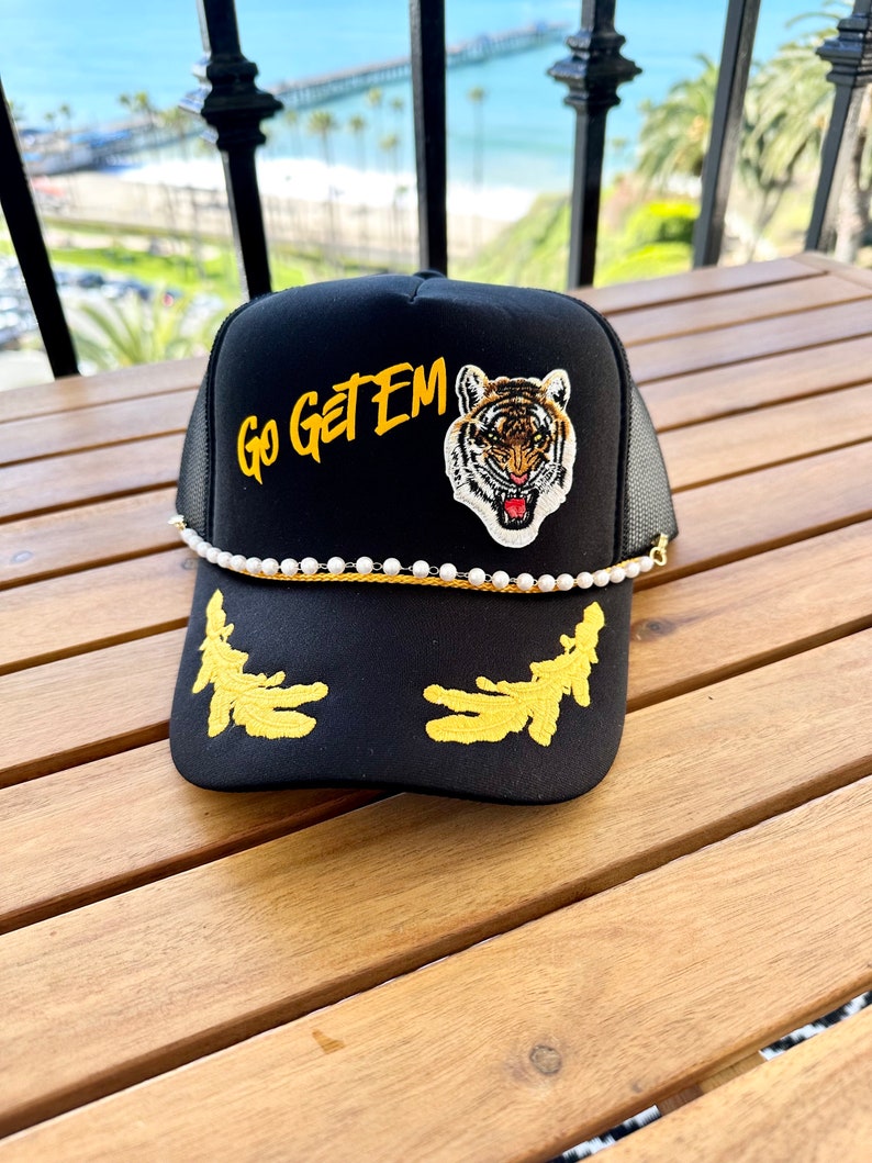 May include: Black trucker hat with a gold embroidered tiger and the words "Go Get Em" in yellow. The hat has a white pearl trim and a black mesh back.