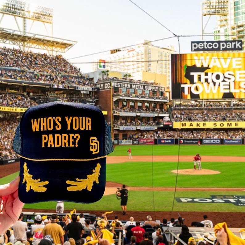 Puede incluir: Una gorra de b&eacute;isbol negra y dorada con el texto "Who's Your Padre?" y el logotipo de los Padres de San Diego. La gorra se sostiene frente a un estadio de b&eacute;isbol.