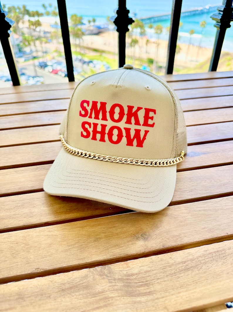 May include: A beige trucker hat with a gold chain trim and red text that reads "SMOKE SHOW".