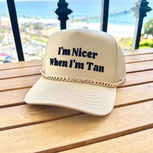 May include: A beige trucker hat with a gold chain around the brim. The hat has black embroidered text that reads "I'm Nicer When I'm Tan."