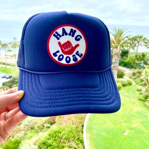 K&ouml;nnte beinhalten: Marineblaue Trucker-Cap mit einem runden Aufn&auml;her in Rot und Wei&szlig; mit der Aufschrift "HANG LOOSE" und einem Handzeichen. Die Kappe hat einen Mesh-R&uuml;cken und einen gebogenen Schirm. Im Hintergrund ist eine K&uuml;stenansicht zu sehen.