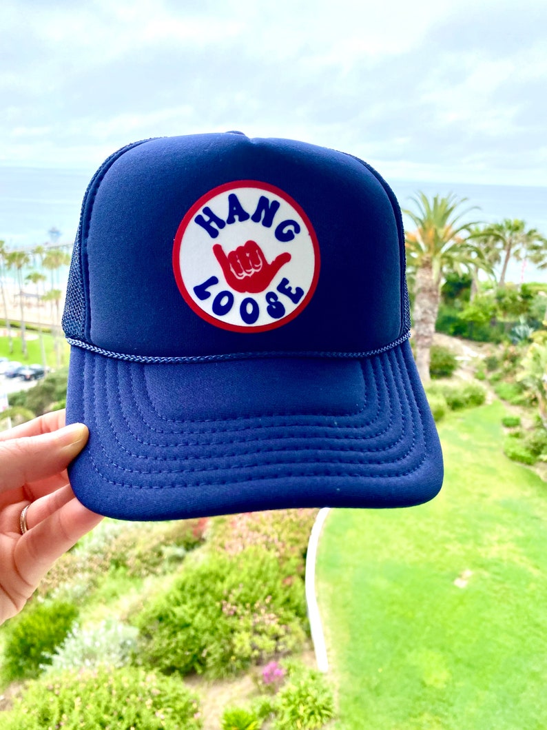 K&ouml;nnte beinhalten: Marineblaue Trucker-Cap mit einem runden Aufn&auml;her in Rot und Wei&szlig; mit der Aufschrift "HANG LOOSE" und einem Handzeichen. Die Kappe hat einen Mesh-R&uuml;cken und einen gebogenen Schirm. Im Hintergrund ist eine K&uuml;stenansicht zu sehen.