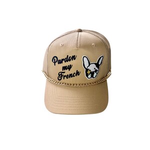 May include: A beige trucker hat with a gold chain trim. The hat has a black embroidered patch with a French bulldog and the text "Pardon my French".