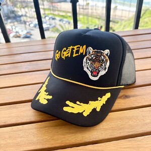 May include: Black trucker hat with a yellow embroidered tiger and the words "Go Get Em" in yellow.
