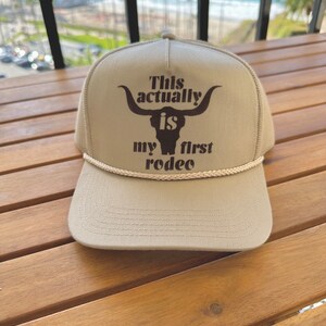 May include: A beige baseball cap with a brown bull's head graphic and the text "This actually is my first rodeo".