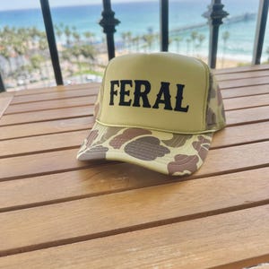 Puede incluir: Una gorra de camionero de camuflaje amarillo y marrón con una malla negra en la parte posterior. La gorra tiene la palabra "FERAL" en letras negras en la parte delantera.